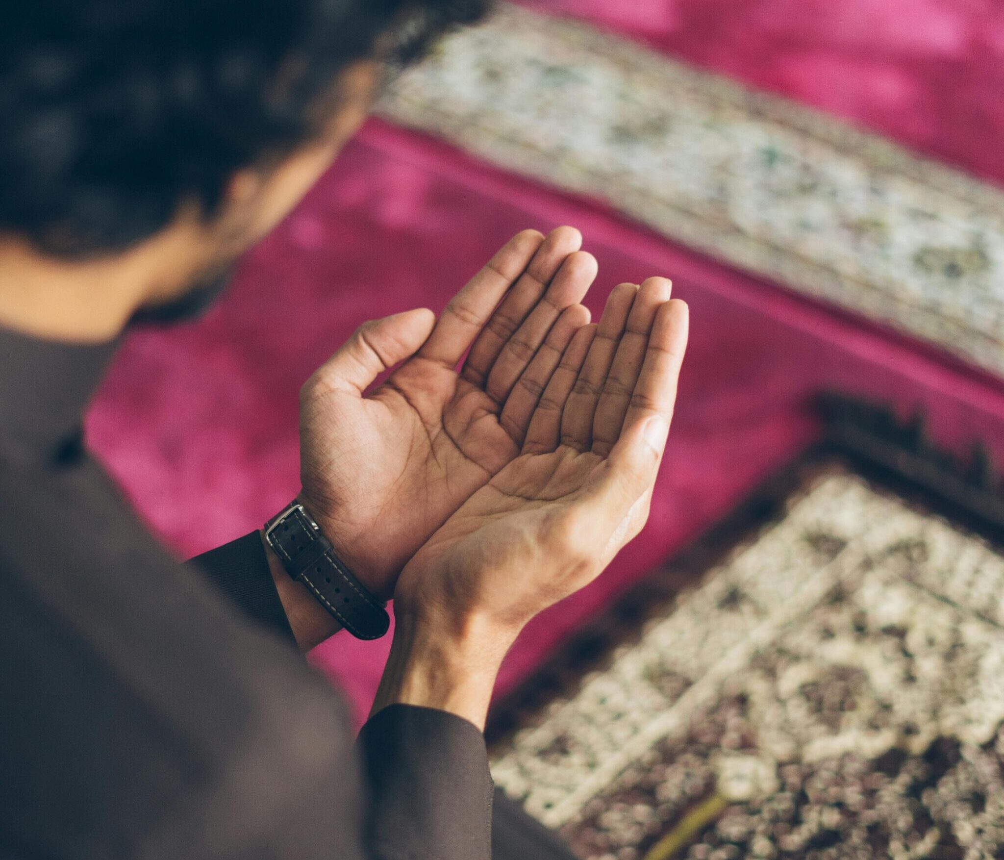 A man in traditional clothing praying indoors on a prayer mat, focusing on his hands lifted in supplication.