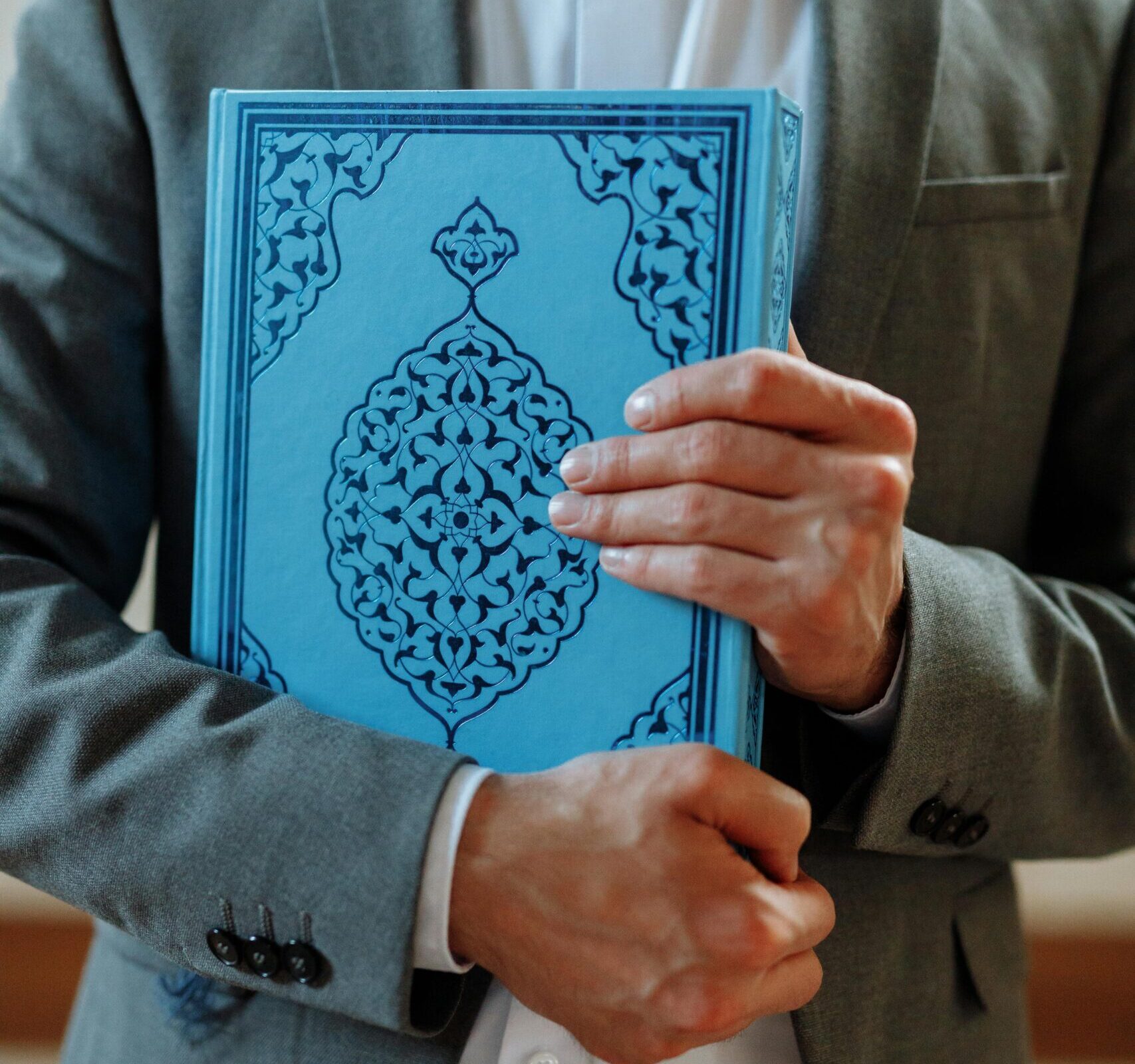 A person in a gray suit holds a blue Islamic book, symbolizing faith.